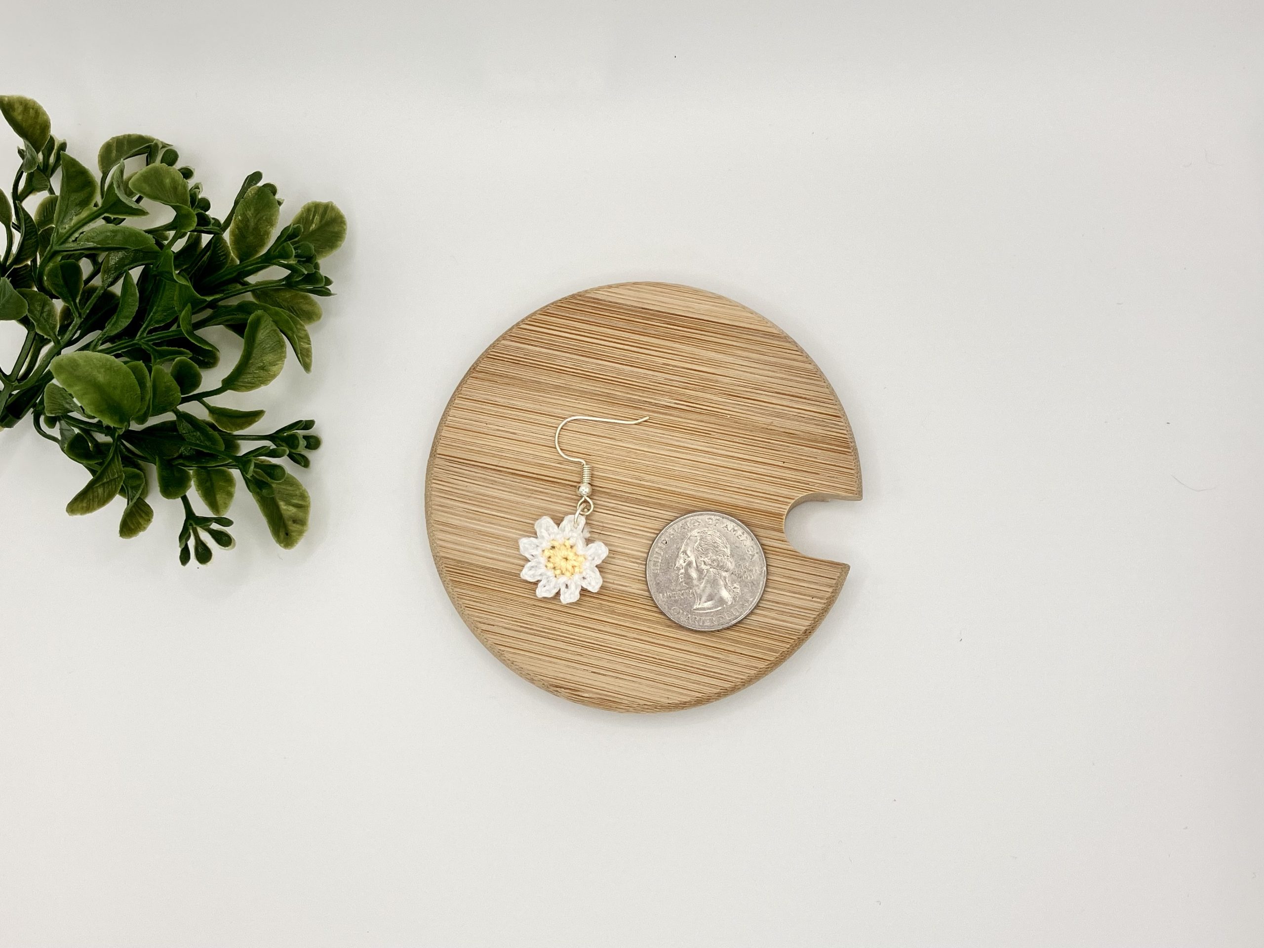 Size comparison of crochet daisy earrings next to a quarter laying flat on a wood coaster