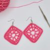 Crochet pink diamond shaped earrings laying next a pink ball of yarn and a metal crochet hook