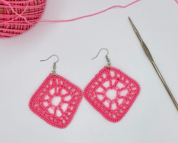 Crochet pink diamond shaped earrings laying next a pink ball of yarn and a metal crochet hook