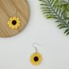 Crochet sunflower earrings, on is up on a wood coaster, the other is further in the right corner laying flat on a white backdrop