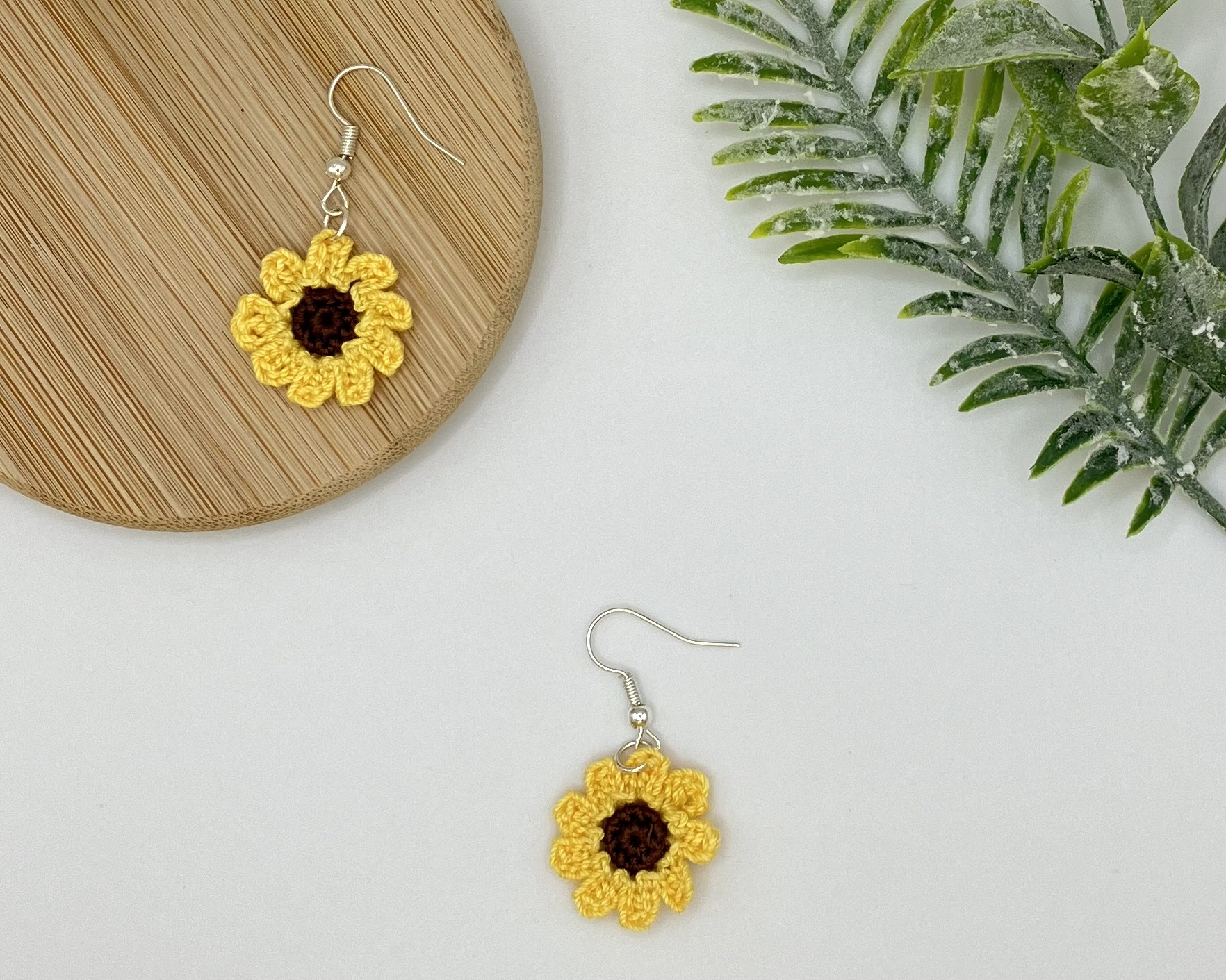 Crochet sunflower earrings, on is up on a wood coaster, the other is further in the right corner laying flat on a white backdrop
