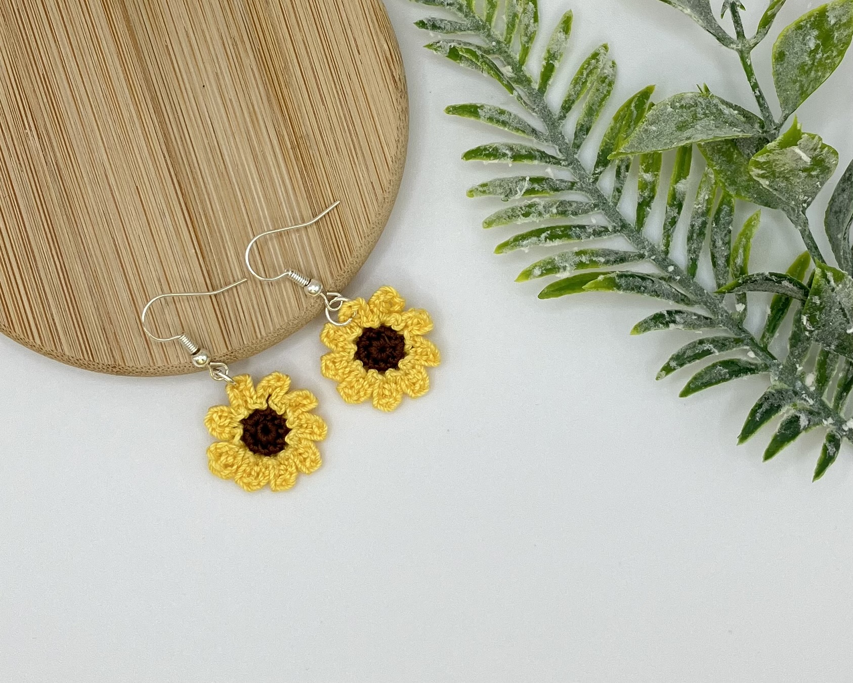 Crochet sunflower earrings with the silver earrings hooks resting on a wood backdrop, and the flower part on white backdrop