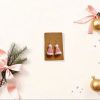 Flat lay of handmade pink Santa hat crochet earrings displayed on a white backdrop, surrounded by minimalistic Christmas props like gold ornaments and sprigs of evergreen, pastel pink ribbon, perfect for holiday gifting.