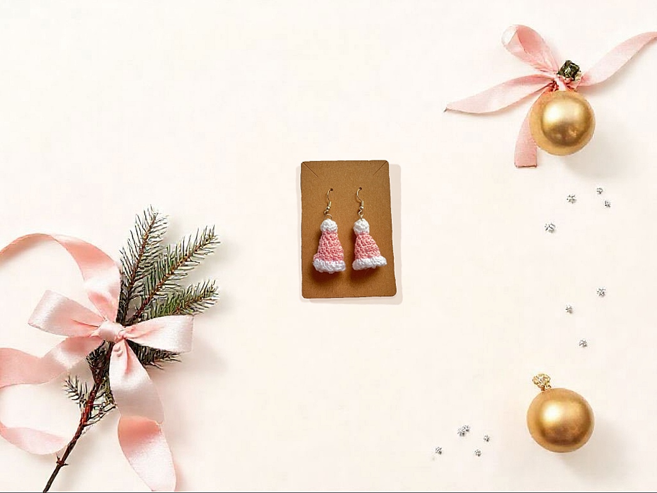 Flat lay of handmade pink Santa hat crochet earrings displayed on a white backdrop, surrounded by minimalistic Christmas props like gold ornaments and sprigs of evergreen, pastel pink ribbon, perfect for holiday gifting.
