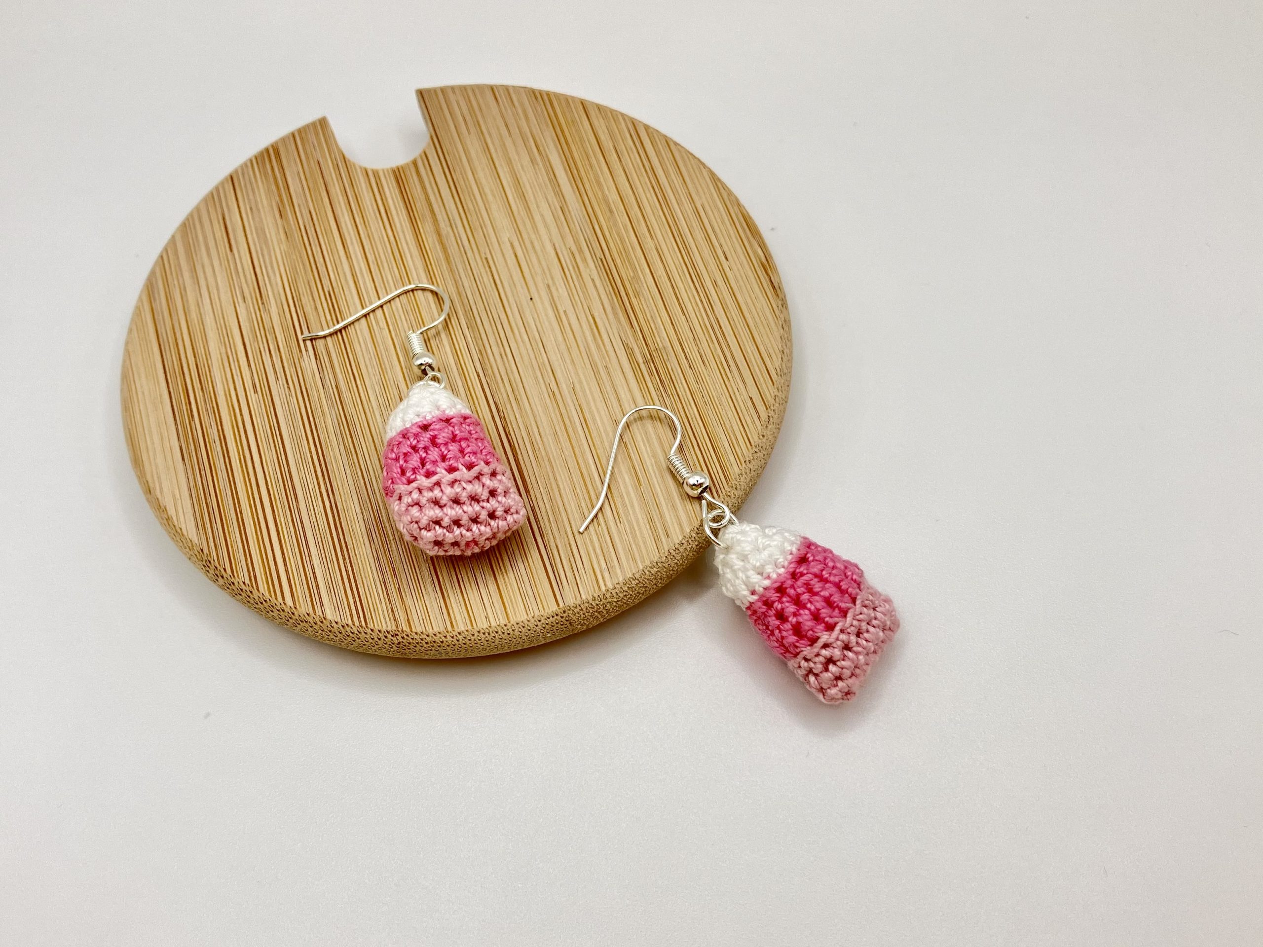 Crocheted pink candy corn earrings one laying on a wood coaster and one on a white backdrop
