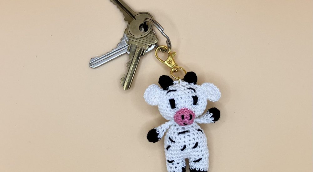 Handmade crochet cow keychain on a soft pastel backdrop with a set of keys