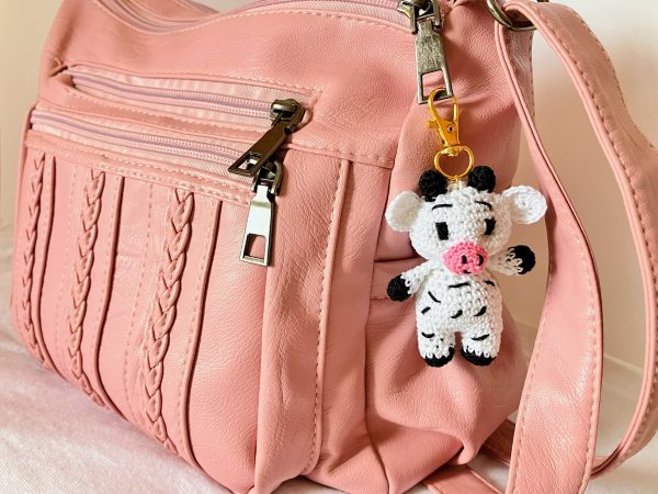 Handmade crochet cow bag charm attached to a pink purse, featuring black and white cotton yarn, perfect for adding a cute accessory to everyday bags.