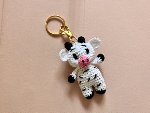 Handmade crochet cow keychain on soft pink backdrop, featuring black and white cotton yarn, ideal for animal-themed accessories.