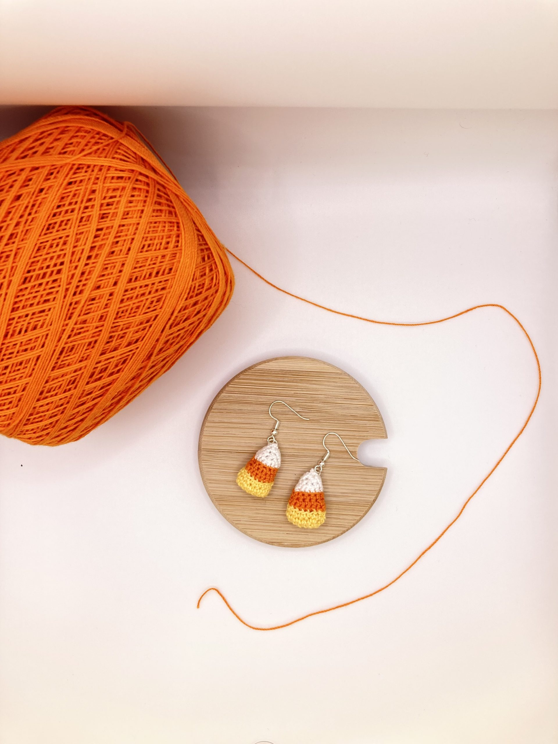 Candy corn earrings laying on a wood coaster next to a ball of orange yarn
