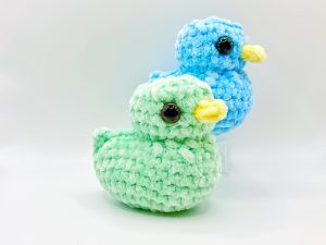 Crochet duck plushie, a green duck in the front, and a blue duck in the back slightly elevated